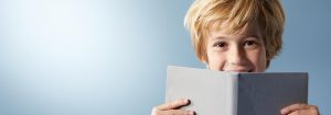 "Jeune enfant aux cheveux blonds souriant derrière un livre ouvert, mettant en valeur une expérience de lecture joyeuse sur fond bleu clair. »