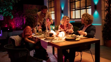 Un groupe d'amis profite d'un dîner en plein air éclairé par une lampe d'extérieur connectée et colorée, disponible avec les offres sur les éclairages d'extérieur connectés. 