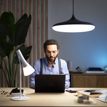 Een man die achter een laptop zit, die op een bureau staat dat wordt verlicht met een slimme tafellamp en een hanglamp met een koelwitte tint licht.