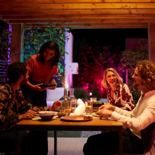 Een groep vrienden geniet samen van een maaltijd op een veranda, die is versierd met kleurrijk slim licht.
