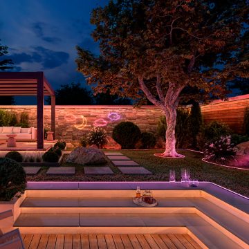 Een achtertuin en patio verlicht met zachte tinten geel, roze en paars licht van verborgen stripverlichting onder trappen en zichtbare stripverlichting langs bloembedden en op muren. 