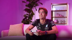 F1 Driver George Russell is sitting on a couch and is holding a video game remote controller in a room with pink lighting.
