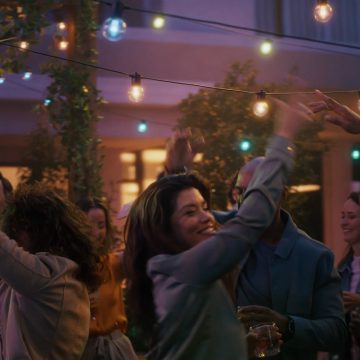 Une femme danse sur un patio sous des guirlandes de lumières de fête à globes Festavia d’extérieur qui projettent un éclairage intelligent bleu, rose et blanc.