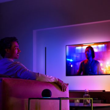 Homme sur un fauteuil qui regarde la télévision avec un éclairage immersif et un haut-parleur intelligent sur la table à côté de lui