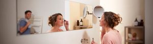 Person applying makeup at a bathroom mirror with bright lighting supporting detailed grooming, bottles and shelves in the background.