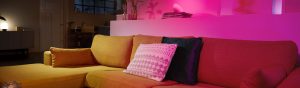 White smart lamp placed on a shelf above a sofa, casting pink light onto the wall and surrounding area in a living room.
