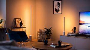Two slim vertical smart lamps placed on the floor and cabinet emit warm light onto the living room walls near furniture and decor.