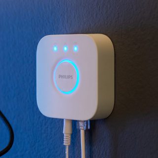 White smart lighting bridge mounted on a blue wall, showing three blue indicator lights and connected via two cables.