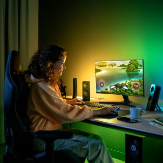 Person gaming at a desk with ambient colored lighting illuminating the wall behind the monitor in a modern home setup.