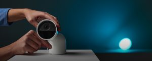 Person adjusting a white smart security camera on a table, with the word ""hue"" visible above the camera lens.
