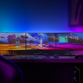 Gaming lights mounted behind monitors cast blue and purple illumination onto the wall above a multi-screen gaming desk setup.