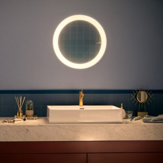 Round illuminated mirror mounted above bathroom sink, emitting bright light onto marble countertop and gold faucet.