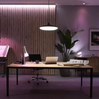 Pendant light fixture mounted above office desk, casting bright light onto workspace with laptop, lamp, and architectural plans.