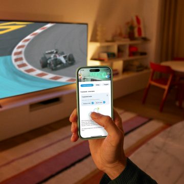 A hand holding a phone using the WiZ App. A TV showing a race lit by WiZ Smart Lighting in the background.