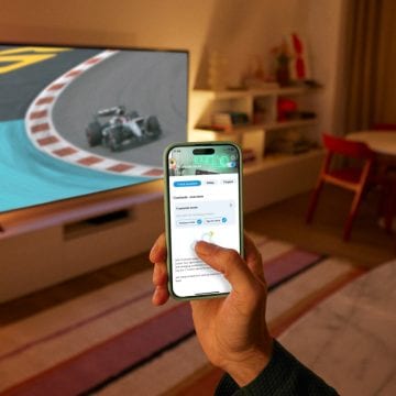 Käsi pitelee puhelinta, jossa on auki WiZ-sovellus. TV:ssä näkyy formulakisat, taustaa valaisee WiZ-älyvalaistus.