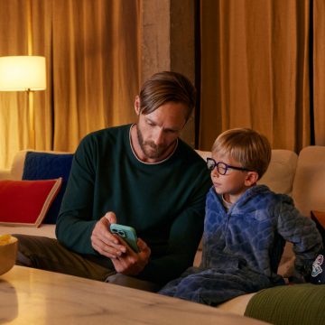 Um pai mostra o telemóvel ao seu filho pequeno no sofá. Um candeeiro com uma lâmpada inteligente WiZ emite uma luz quente.
